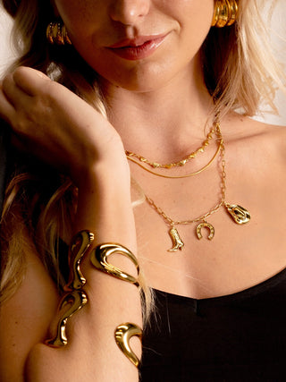 Close-up of a person wearing layered gold necklaces and bold gold cuffs, styled with Western-inspired charms and polished gold earrings against a black top.