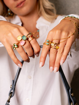 Close-up of hands stacked with gold rings featuring colorful gemstones in green, pink, blue, yellow, and red, paired with gold chain bracelets and a white shirt while holding a leather bag.