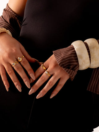 Close-up of hands wearing chunky ivory bangles and gold rings with nude, almond-shaped nails, styled against a dark outfit with striped brown sleeves.