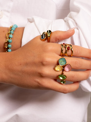 Close-up of a hand wearing multiple colourful gemstone rings in green, blue, yellow, and pink tones, paired with gold statement rings and a turquoise beaded bracelet, styled with a white shirt.