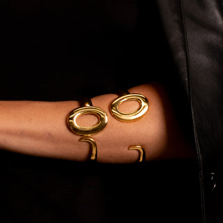Close-up of model wearing two gold Spencer Lock bangles stacked on the forearm, showcasing their sculptural oval design against a black outfit. Modern, bold and luxurious Forward & Tame styling.