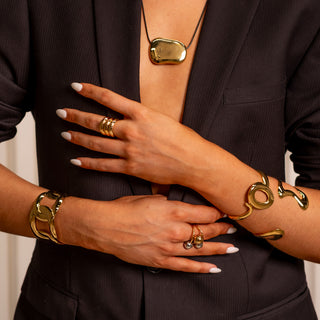 Editorial portrait of model wearing gold sculptural bangles, layered necklaces and hoop earrings. Warm lighting, black outfit and relaxed styling for Forward & Tame’s luxury look.
