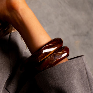 Close up of two Terra resin bangles in warm brown marble effect, worn stacked on the wrist with a grey blazer, emphasising texture, shine and statement styling.